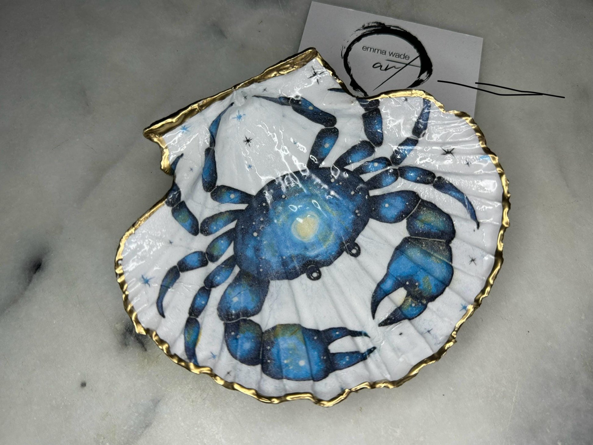 Scallop shell trinket dish with a hand drawn and hand painted blue crab design from original watercolour artwork. The artwork is applied as a decoupage print, then enhanced with painted detailing for a unique finish. Each shell is edged with liquid gold leaf and sealed with glossy resin. A handmade shell ring dish, jewellery organiser, or trinket tray for rings, earrings, or cufflinks. Perfect coastal decor or gift for weddings, anniversaries, birthdays, or Christmas.