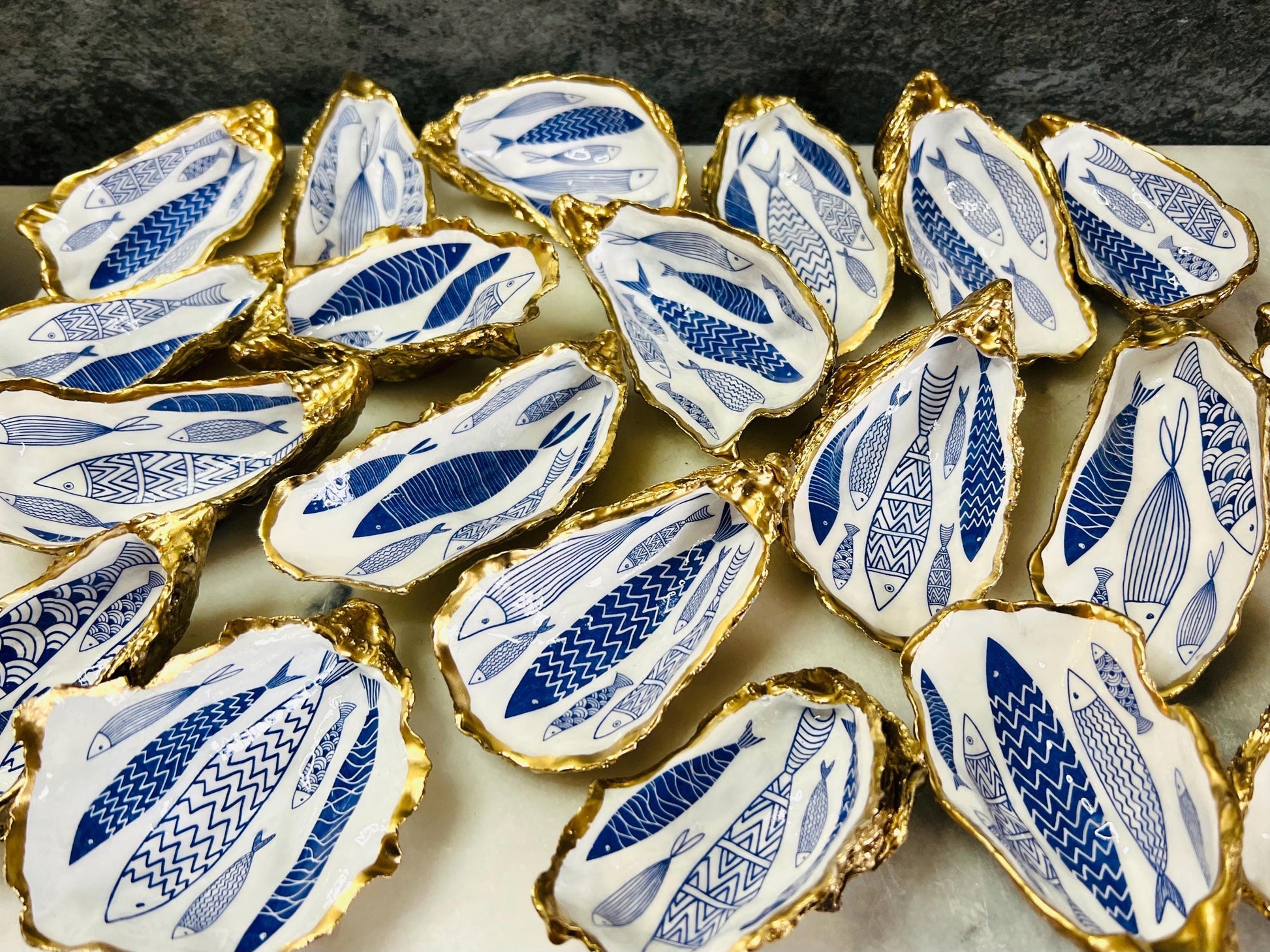 Hand-gilded Dorset oyster shell trinket dish featuring a blue fish motif, edged with liquid gold leaf and sealed in high gloss resin. Coastal ring holder and nautical home decor piece displayed with multiple shells. Sustainable oyster shell art handmade in Surrey, ideal as a jewellery dish, wedding favour, beach house accessory or ocean lover gift.