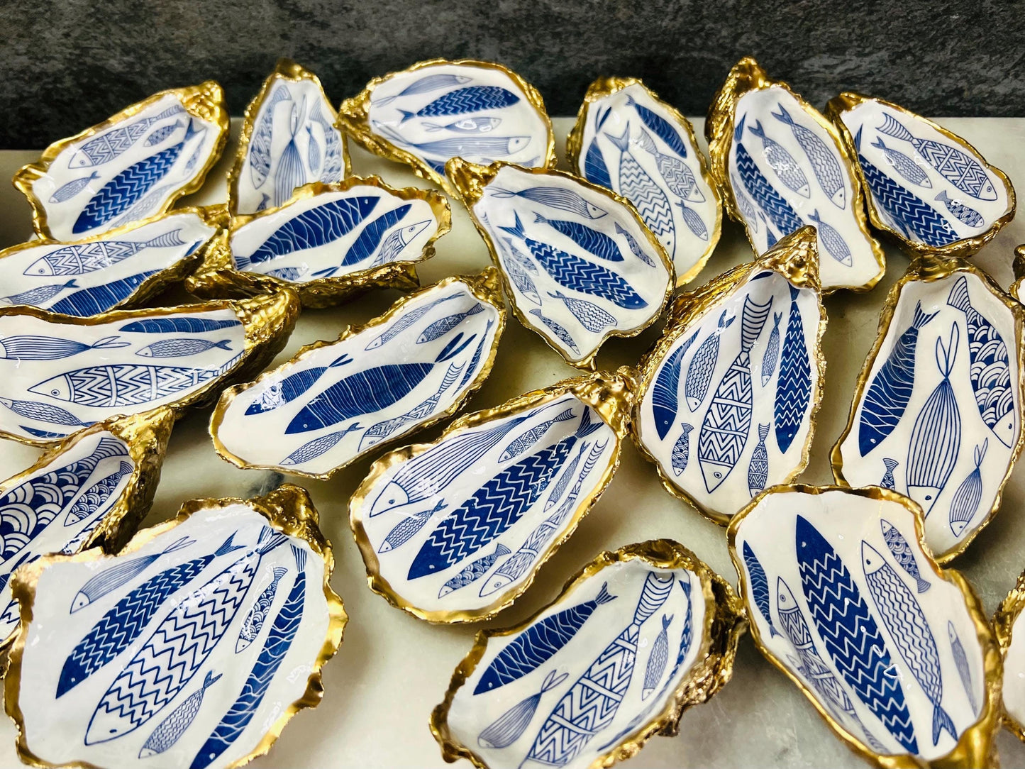 Hand-gilded Dorset oyster shell trinket dish featuring a blue fish motif, edged with liquid gold leaf and sealed in high gloss resin. Coastal ring holder and nautical home decor piece displayed with multiple shells. Sustainable oyster shell art handmade in Surrey, ideal as a jewellery dish, wedding favour, beach house accessory or ocean lover gift.