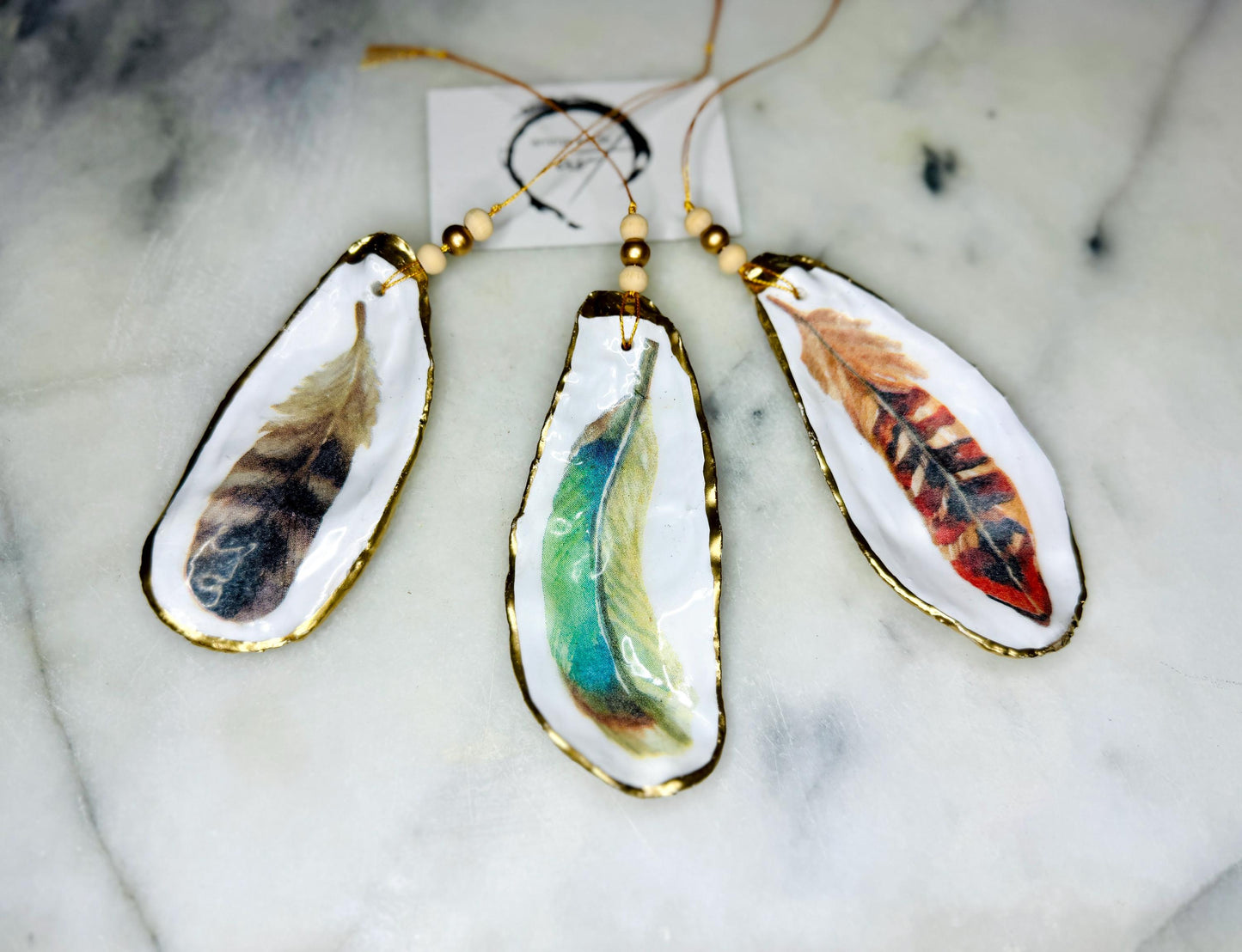 Aquarelle feather oyster shell hanging ornaments, handmade coastal décor, gilded gold leaf shells, resin sealed feather decorations, Christmas tree ornaments, nature-inspired gifts, watercolor feather oyster shells, Dorset handmade shell ornaments, sustainable coastal gifts, oyster shell art, feather design decorations, unique shell ornaments, eco-friendly handmade oyster shell Christmas gifts.