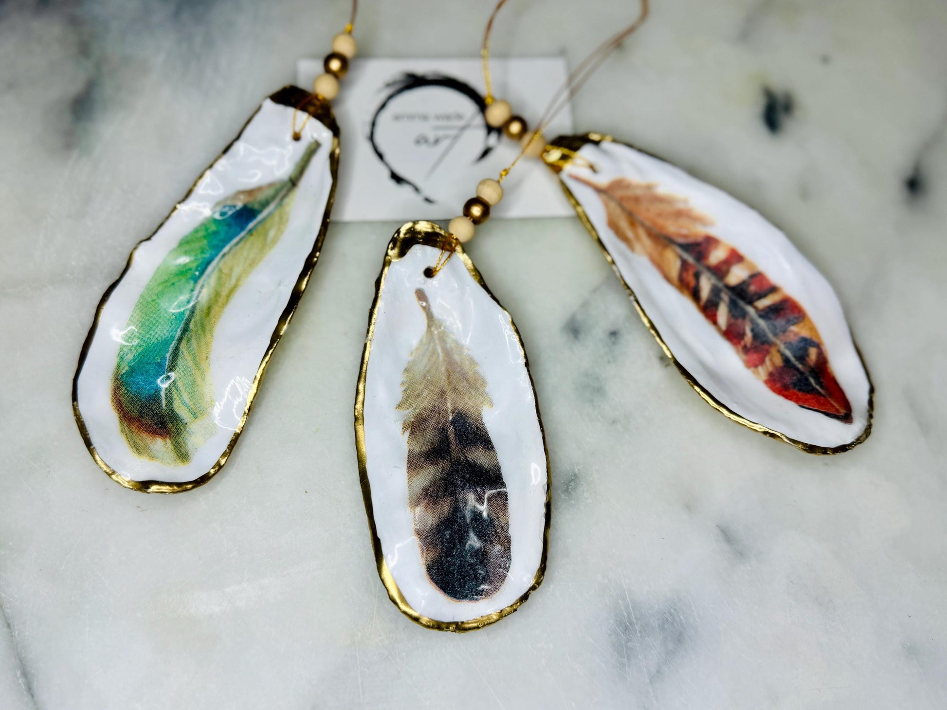 Aquarelle feather oyster shell hanging ornaments, handmade coastal décor, gilded gold leaf shells, resin sealed feather decorations, Christmas tree ornaments, nature-inspired gifts, watercolor feather oyster shells, Dorset handmade shell ornaments, sustainable coastal gifts, oyster shell art, feather design decorations, unique shell ornaments, eco-friendly handmade oyster shell Christmas gifts.