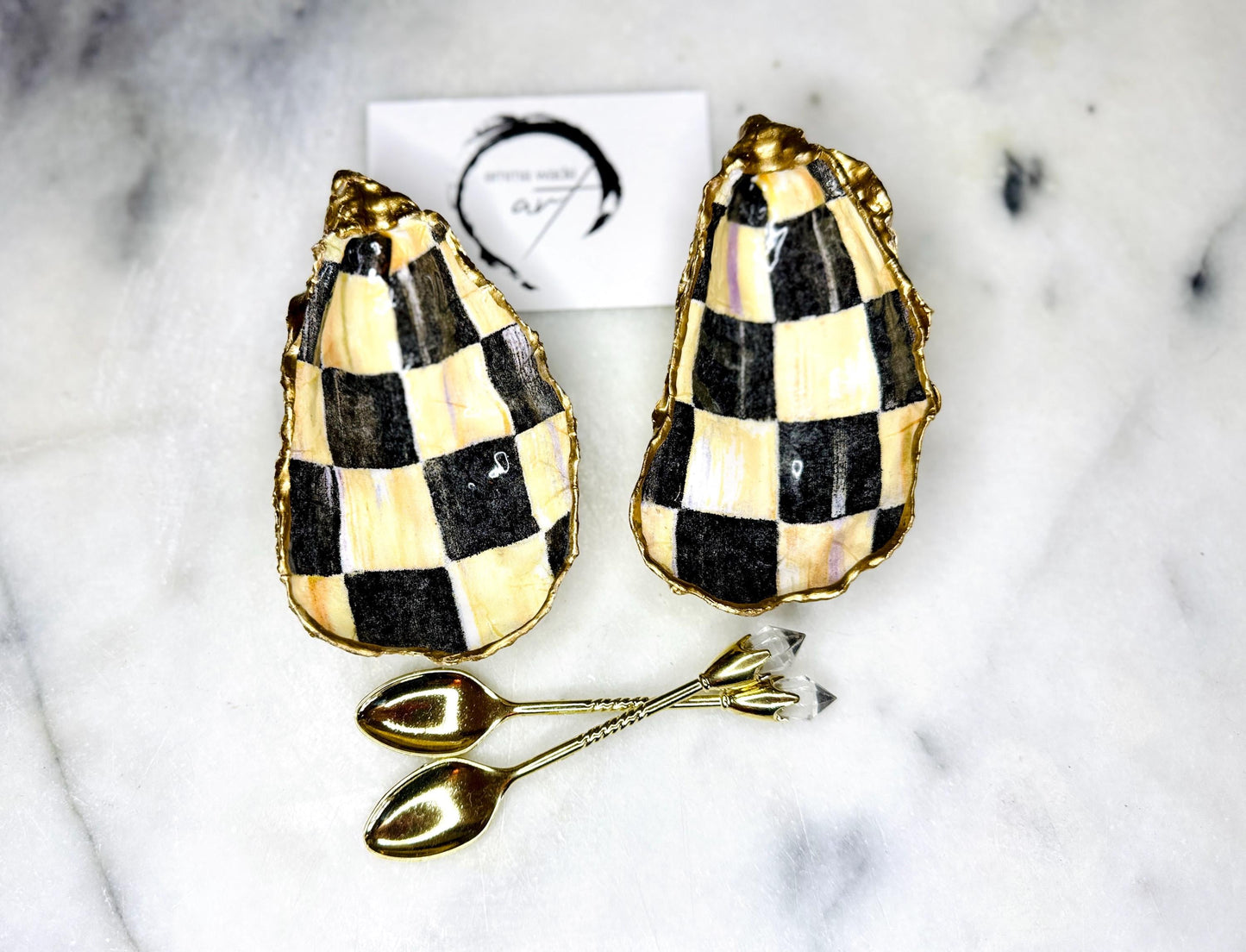 Handmade black, pearl and gold chequered oyster shell salt and pepper pinch pots, shown as a matching pair with small gold spoons on a light background. Natural Dorset shells decorated with a graphic pattern, gilded with gold leaf and sealed with a glossy, food-safe resin. Elegant coastal table decor and a unique gift for weddings, anniversaries or stylish dining tables, each piece one of a kind.