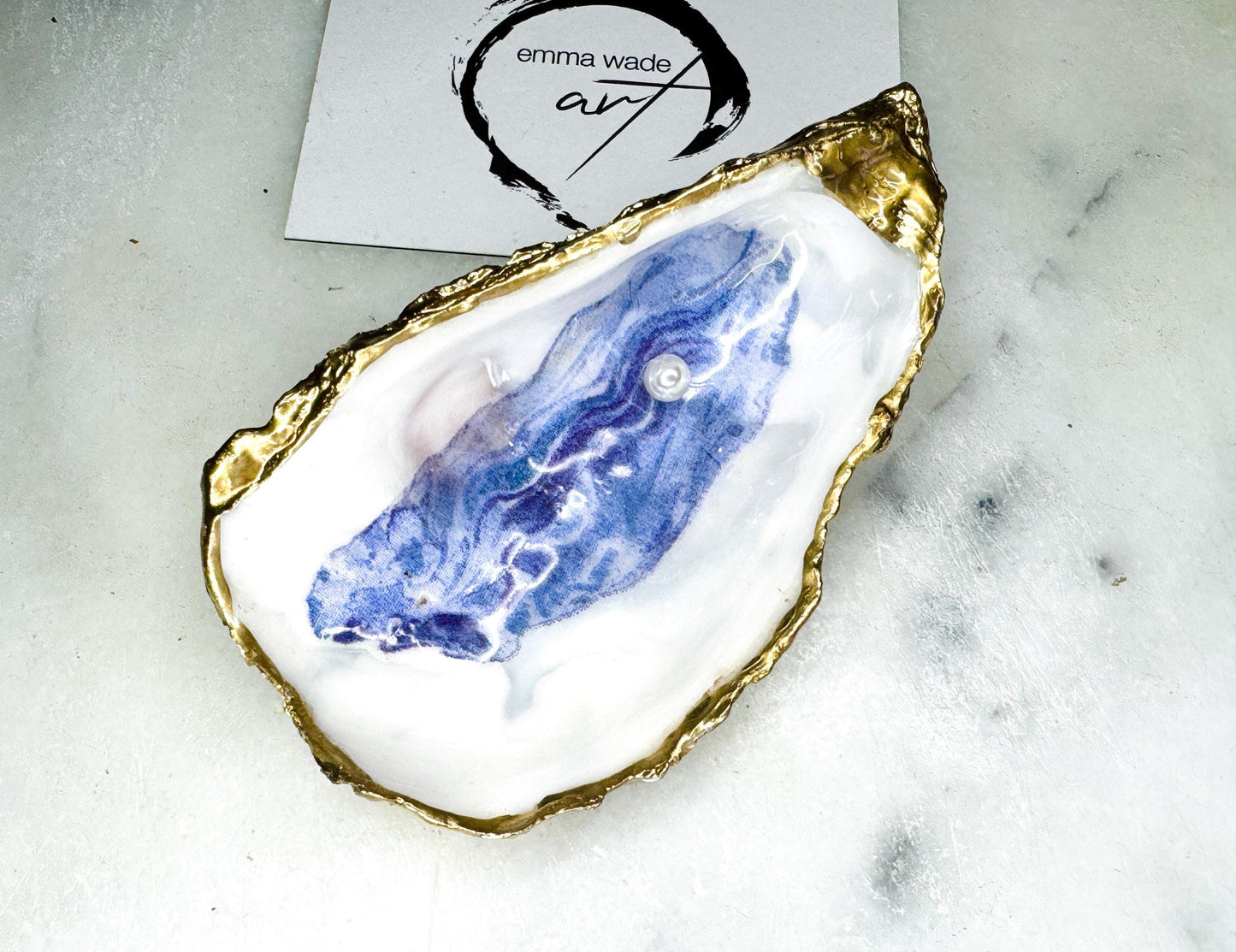 Handmade Dorset oyster shell trinket dish featuring a blue and beige watercolour oyster illustration with Swarovski pearl detail, finished with hand-applied gold leaf edging and glossy resin. Coastal-inspired jewellery dish and ring holder available in multiple sizes including XL oyster shells. Elegant handmade coastal décor gift ideal for weddings, birthdays, anniversaries, and luxury beach house interiors.