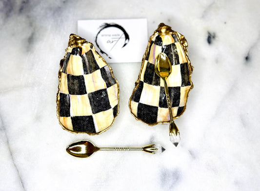 Handmade black, pearl and gold chequered oyster shell salt and pepper pinch pots, shown as a matching pair with small gold spoons on a light background. Natural Dorset shells decorated with a graphic pattern, gilded with gold leaf and sealed with a glossy, food-safe resin. Elegant coastal table decor and a unique gift for weddings, anniversaries or stylish dining tables, each piece one of a kind.