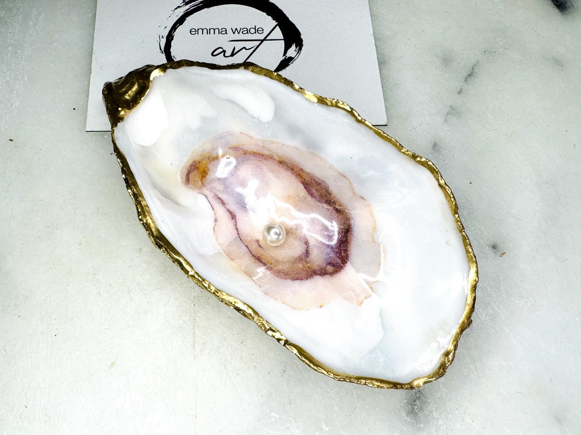 Handmade Dorset oyster shell trinket dish featuring a blue and beige watercolour oyster illustration with Swarovski pearl detail, finished with hand-applied gold leaf edging and glossy resin. Coastal-inspired jewellery dish and ring holder available in multiple sizes including XL oyster shells. Elegant handmade coastal décor gift ideal for weddings, birthdays, anniversaries, and luxury beach house interiors.