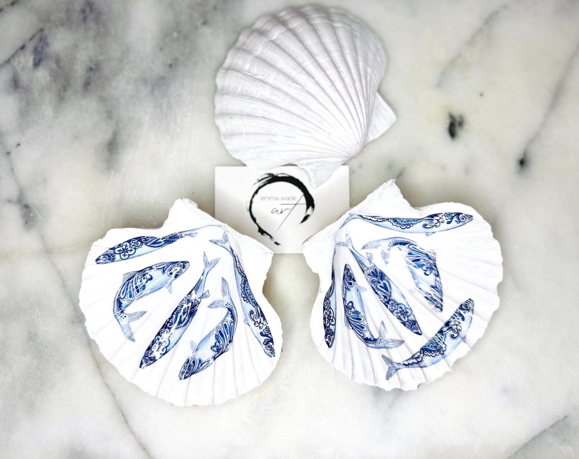 Handmade Dorset scallop shell trinket dishes featuring blue and white paisley fish illustrations with hand-painted gold leaf edging and glossy crystal clear resin. Coastal Scandinavian inspired jewellery dishes styled on a soft neutral background with elegant nautical fish artwork and luxurious gilded detailing.