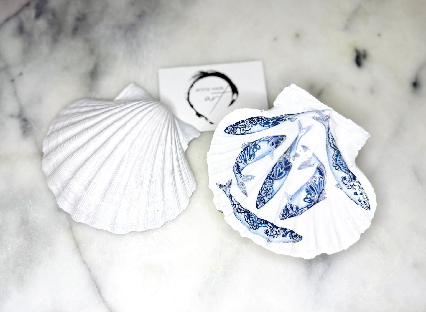 Handmade Dorset scallop shell trinket dishes featuring blue and white paisley fish illustrations with hand-painted gold leaf edging and glossy crystal clear resin. Coastal Scandinavian inspired jewellery dishes styled on a soft neutral background with elegant nautical fish artwork and luxurious gilded detailing.