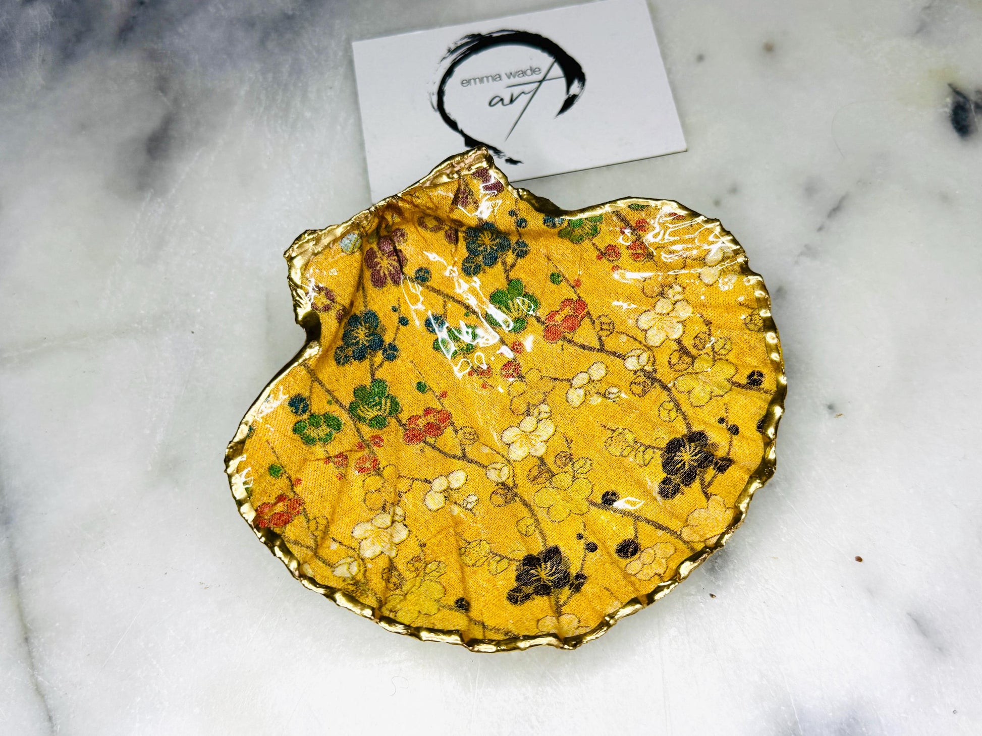 Handcrafted Dorset scallop shell trinket dish featuring a Japanese plum blossom floral design on a rich yellow background, finished with a gold leaf rim and glossy resin. Decorative blossom branches in white, green and black tones, styled on a light marble surface with a gold shell accent. Coastal inspired jewellery tray or ring holder, ideal as a handmade decorative keepsake gift.