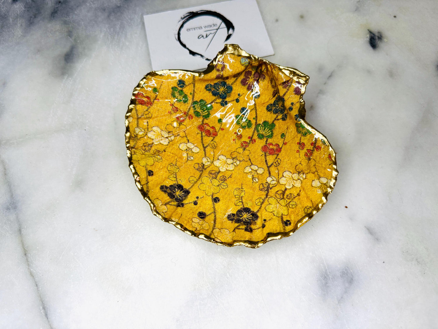 Handcrafted Dorset scallop shell trinket dish featuring a Japanese plum blossom floral design on a rich yellow background, finished with a gold leaf rim and glossy resin. Decorative blossom branches in white, green and black tones, styled on a light marble surface with a gold shell accent. Coastal inspired jewellery tray or ring holder, ideal as a handmade decorative keepsake gift.