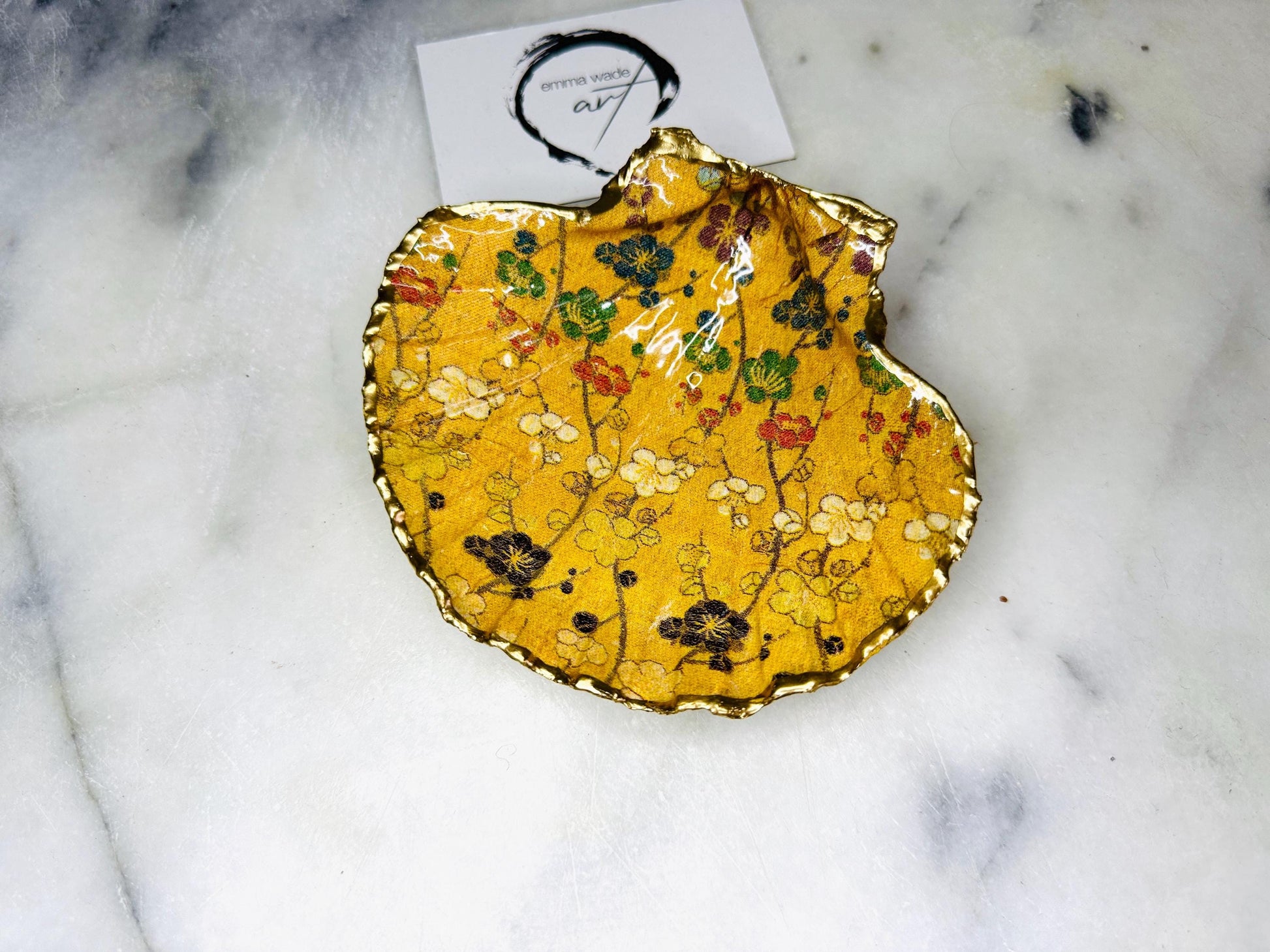 Handcrafted Dorset scallop shell trinket dish featuring a Japanese plum blossom floral design on a rich yellow background, finished with a gold leaf rim and glossy resin. Decorative blossom branches in white, green and black tones, styled on a light marble surface with a gold shell accent. Coastal inspired jewellery tray or ring holder, ideal as a handmade decorative keepsake gift.