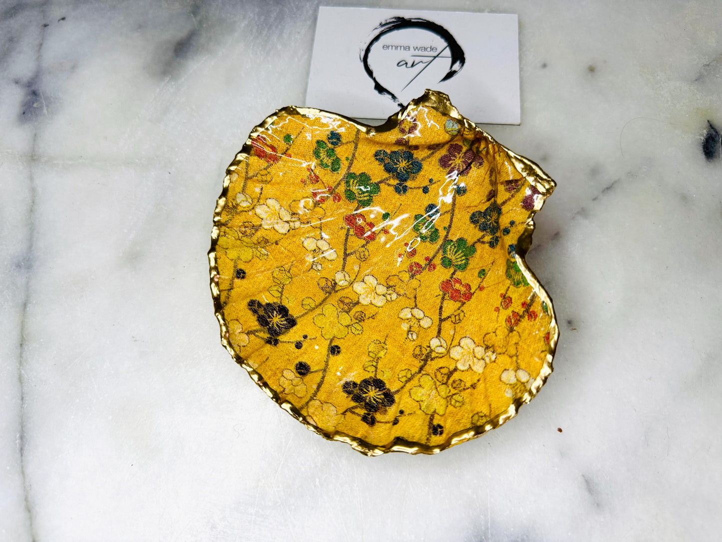 Handcrafted Dorset scallop shell trinket dish featuring a Japanese plum blossom floral design on a rich yellow background, finished with a gold leaf rim and glossy resin. Decorative blossom branches in white, green and black tones, styled on a light marble surface with a gold shell accent. Coastal inspired jewellery tray or ring holder, ideal as a handmade decorative keepsake gift.