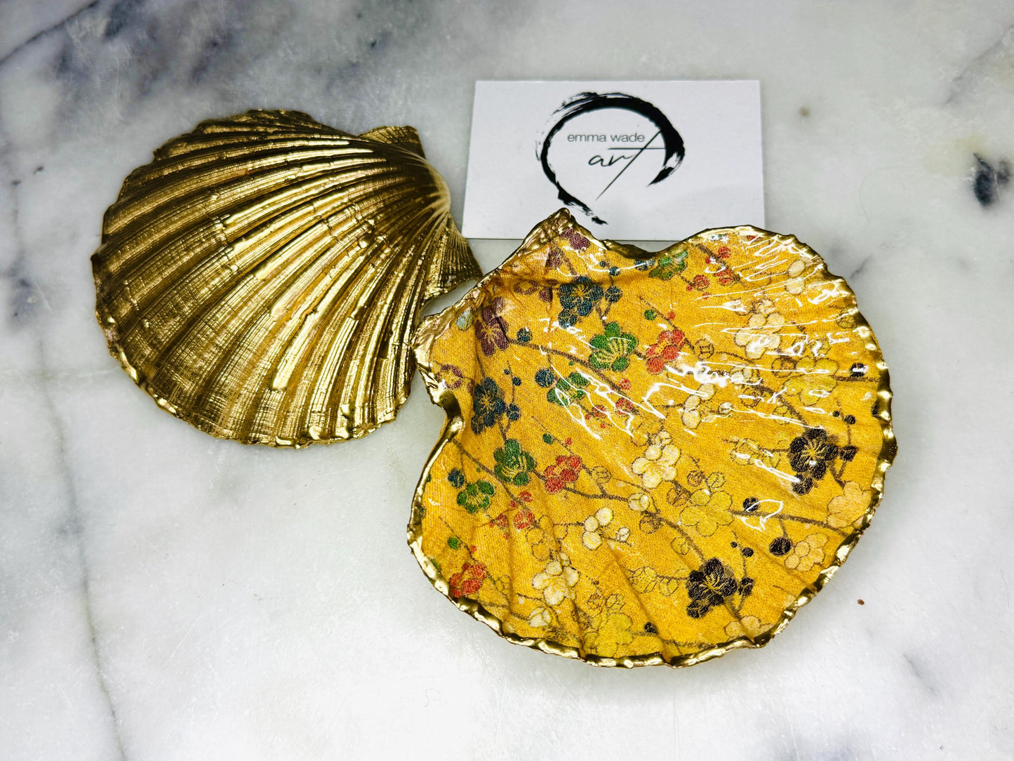 Handcrafted Dorset scallop shell trinket dish featuring a Japanese plum blossom floral design on a rich yellow background, finished with a gold leaf rim and glossy resin. Decorative blossom branches in white, green and black tones, styled on a light marble surface with a gold shell accent. Coastal inspired jewellery tray or ring holder, ideal as a handmade decorative keepsake gift.