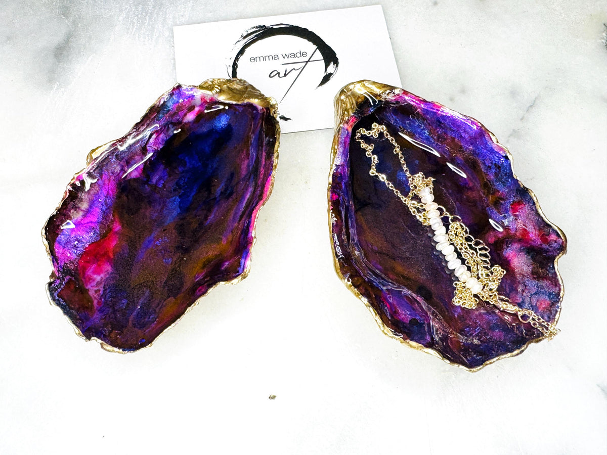 Handmade Dorset oyster shell salt and pepper pinch pot set featuring dramatic purple, blue and magenta “sea cave” interiors with hand-painted liquid gold leaf edging. Coastal ocean-inspired shell dishes with glossy resin finish and matching gold crystal spoons, styled on a white marble background.