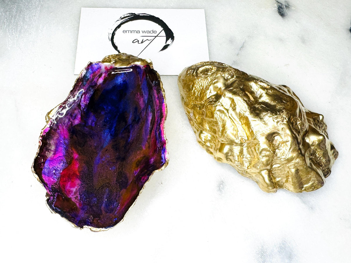 Handmade Dorset oyster shell salt and pepper pinch pot set featuring dramatic purple, blue and magenta “sea cave” interiors with hand-painted liquid gold leaf edging. Coastal ocean-inspired shell dishes with glossy resin finish and matching gold crystal spoons, styled on a white marble background.