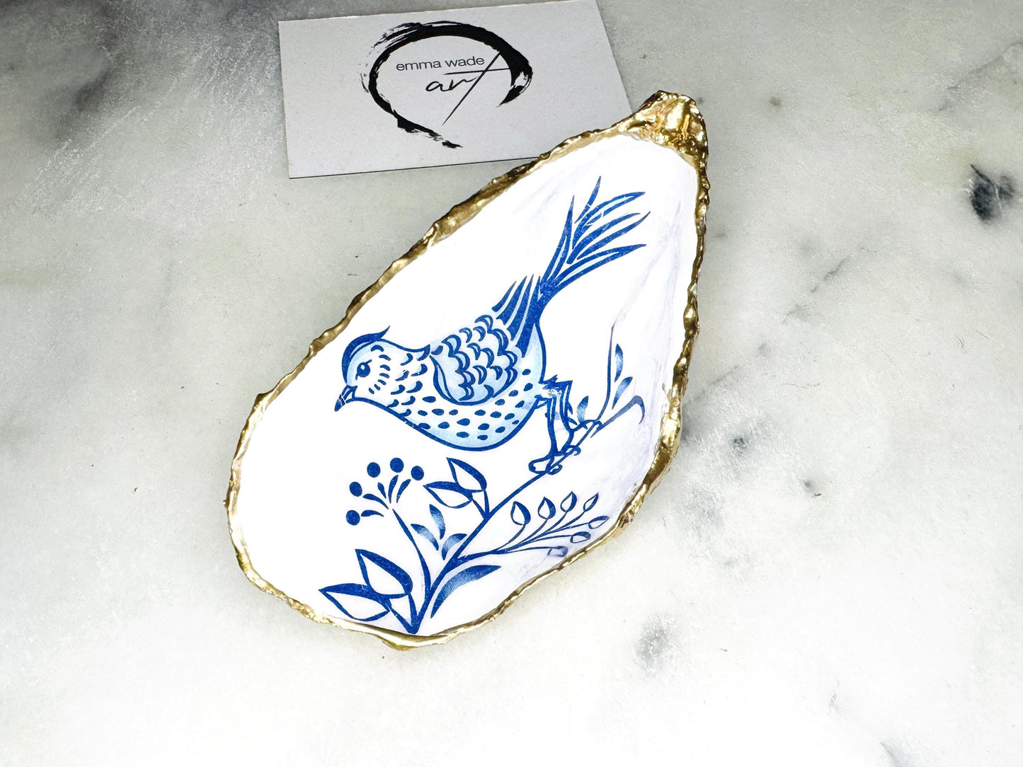 Handmade Dorset oyster shell trinket dish with blue porcelain-style bird design, gold leaf gilded edges and glossy resin finish. Coastal jewellery holder and ring dish photographed on a light surface, showing the natural shell shape and pearly interior. Unique sustainably sourced shell, sold individually as decorative coastal home décor or a thoughtful gift.