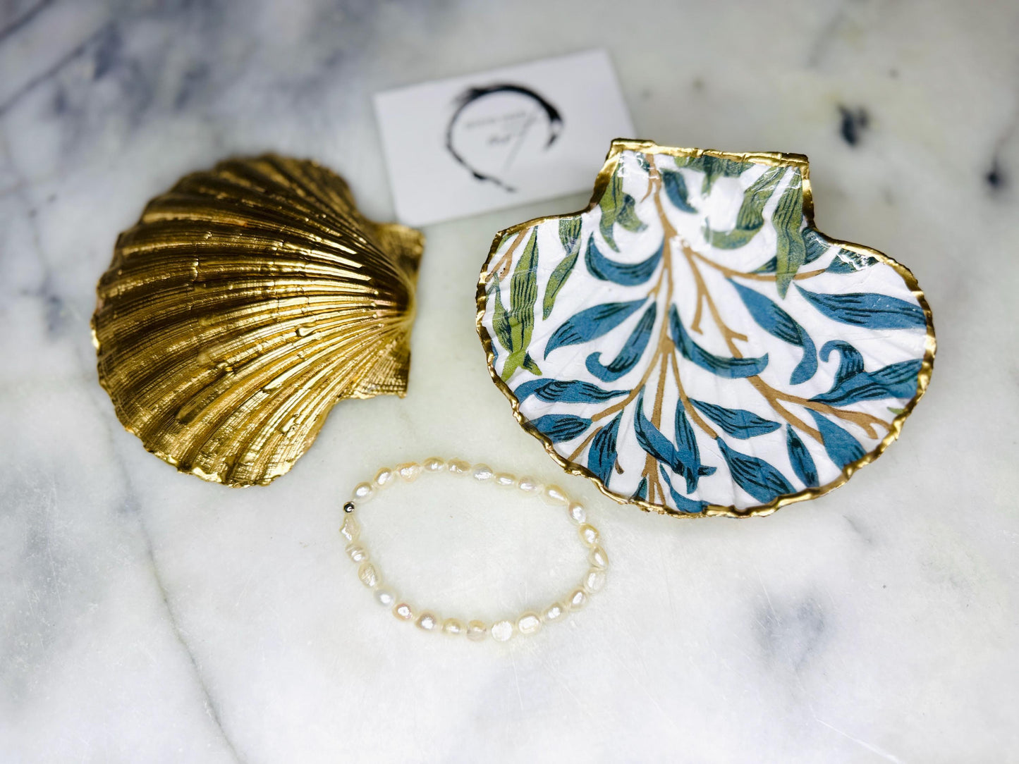 Handmade Dorset scallop shell trinket dish featuring William Morris Willow Bough design in soft blue and green tones. Decorative shell jewellery dish finished with hand-applied liquid gold leaf edging and glossy resin. Coastal inspired ring holder with botanical Arts and Crafts pattern and natural shell shape, styled as elegant bathroom décor and heritage home accessory.