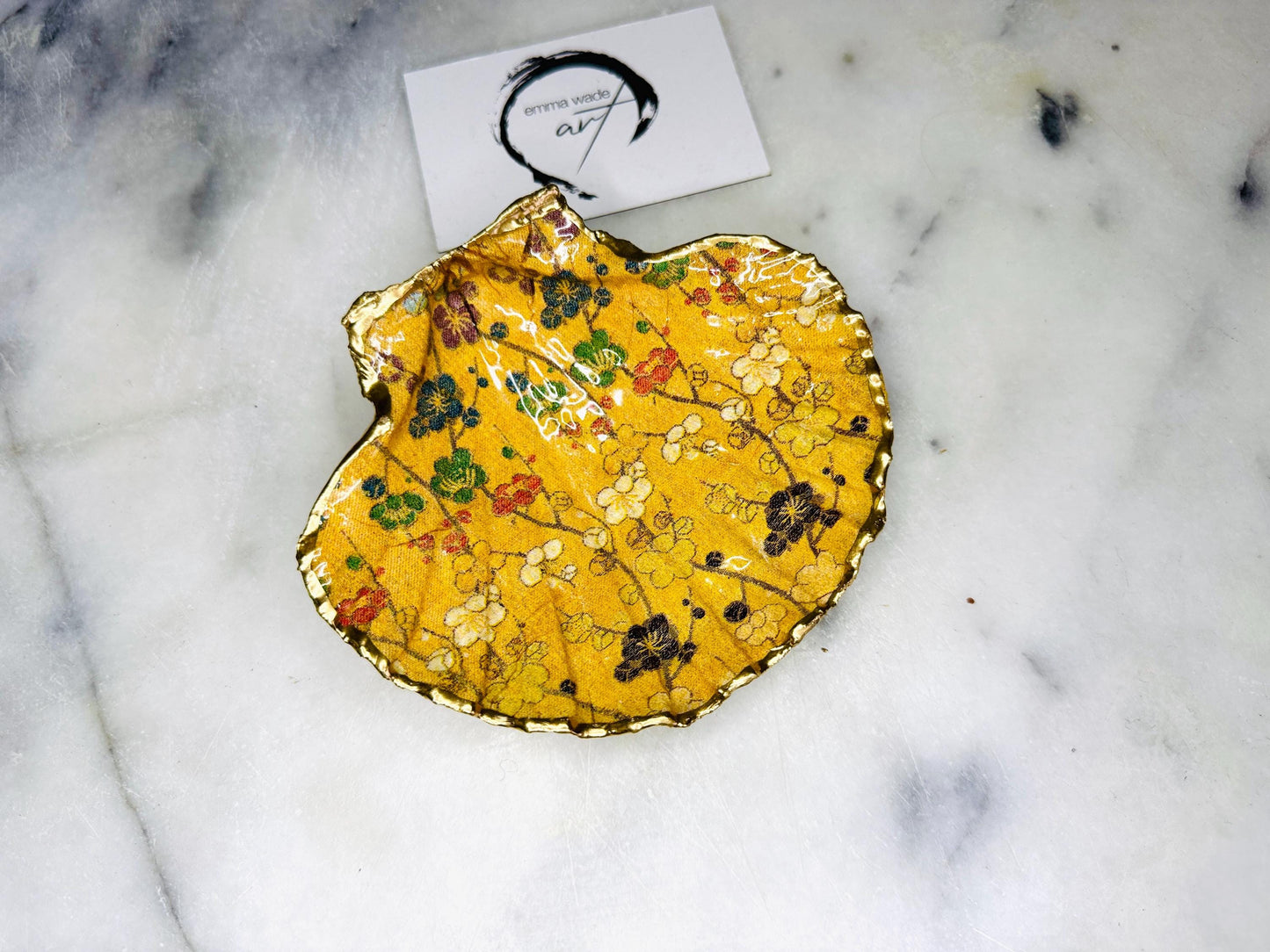 Handcrafted Dorset scallop shell trinket dish featuring a Japanese plum blossom floral design on a rich yellow background, finished with a gold leaf rim and glossy resin. Decorative blossom branches in white, green and black tones, styled on a light marble surface with a gold shell accent. Coastal inspired jewellery tray or ring holder, ideal as a handmade decorative keepsake gift.