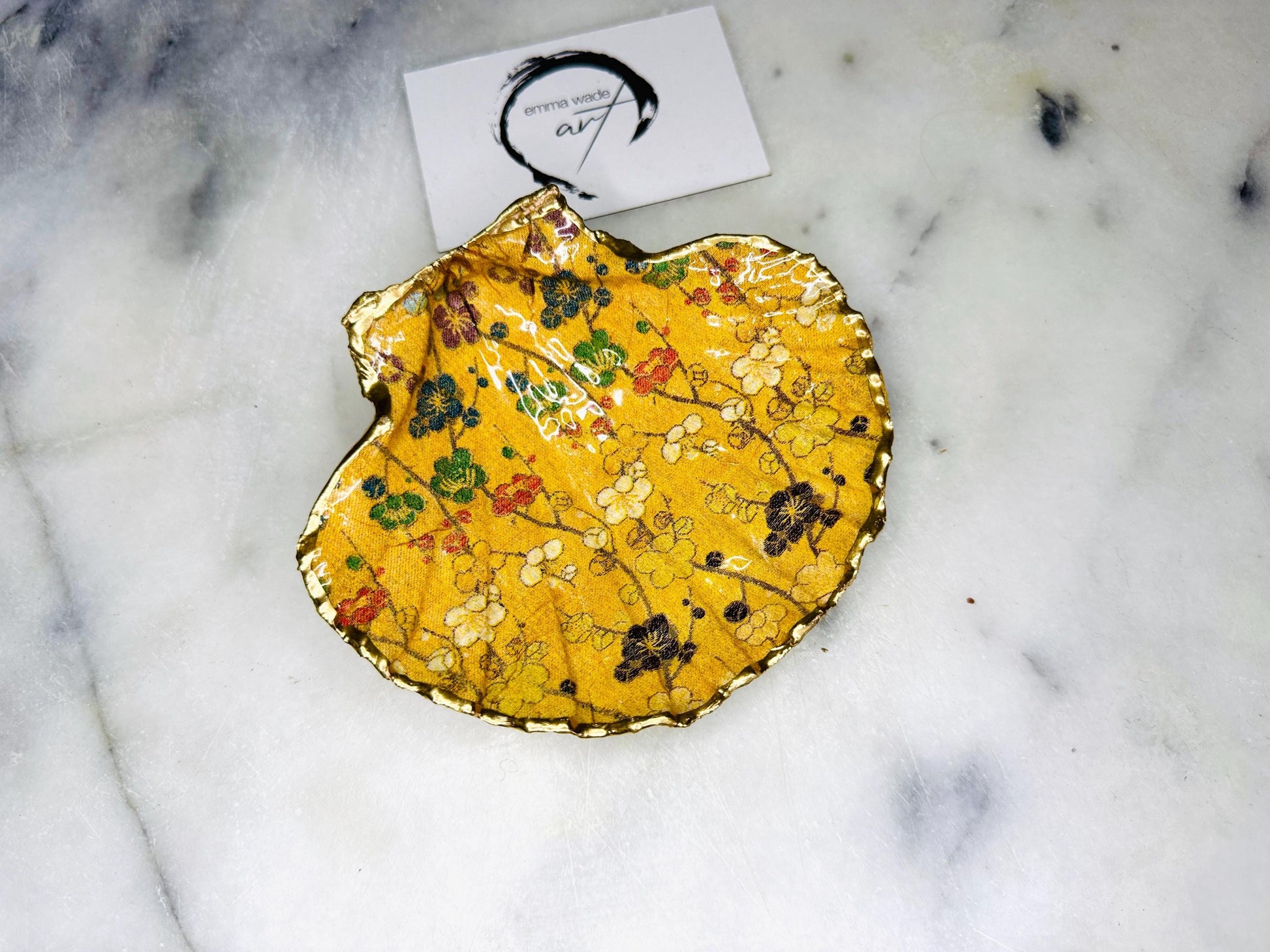 Handcrafted Dorset scallop shell trinket dish featuring a Japanese plum blossom floral design on a rich yellow background, finished with a gold leaf rim and glossy resin. Decorative blossom branches in white, green and black tones, styled on a light marble surface with a gold shell accent. Coastal inspired jewellery tray or ring holder, ideal as a handmade decorative keepsake gift.