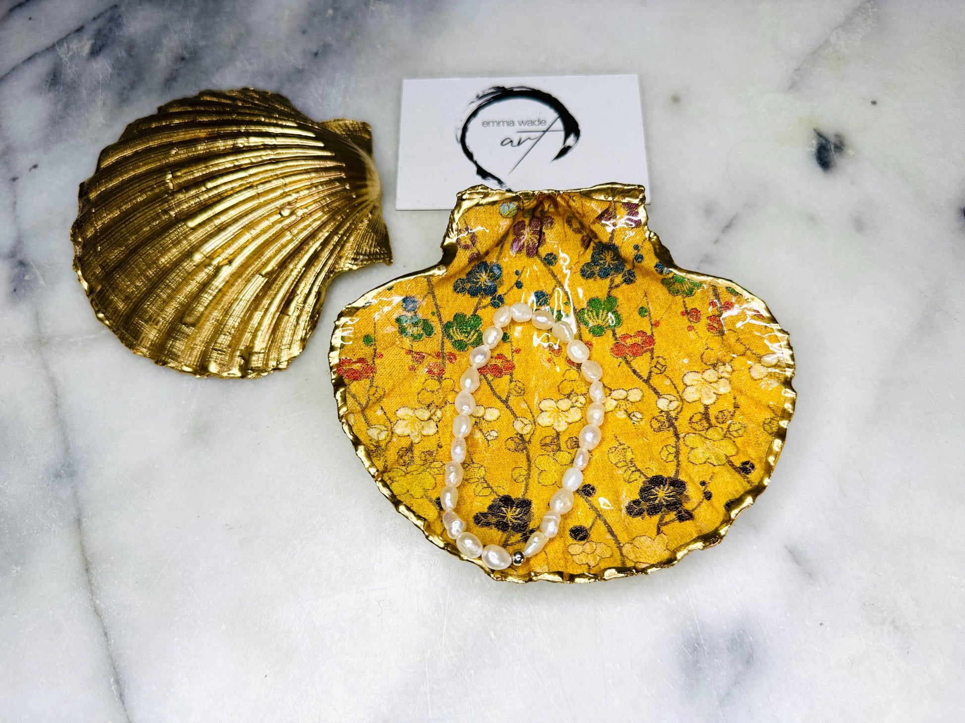 Handcrafted Dorset scallop shell trinket dish featuring a Japanese plum blossom floral design on a rich yellow background, finished with a gold leaf rim and glossy resin. Decorative blossom branches in white, green and black tones, styled on a light marble surface with a gold shell accent. Coastal inspired jewellery tray or ring holder, ideal as a handmade decorative keepsake gift.