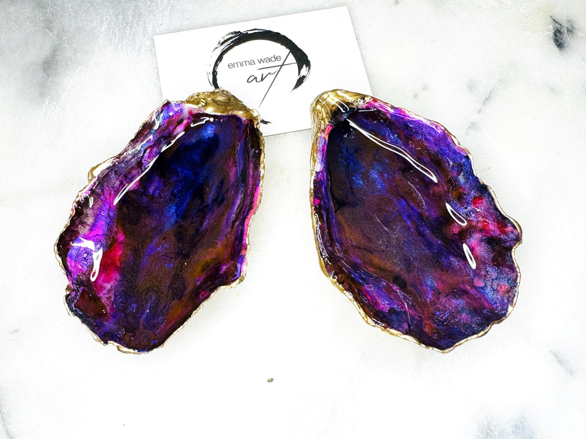 Handmade Dorset oyster shell salt and pepper pinch pot set featuring dramatic purple, blue and magenta “sea cave” interiors with hand-painted liquid gold leaf edging. Coastal ocean-inspired shell dishes with glossy resin finish and matching gold crystal spoons, styled on a white marble background.