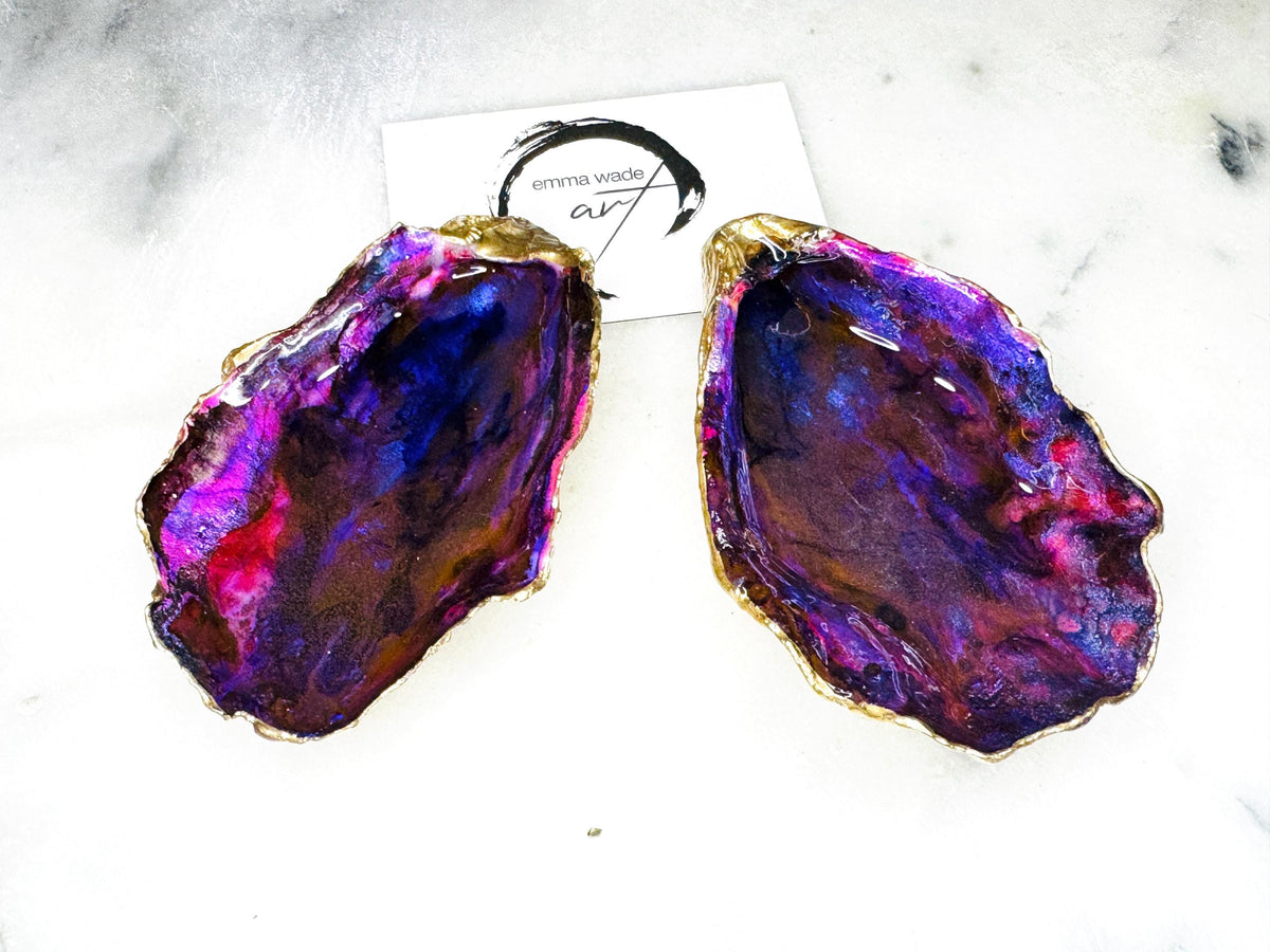 Handmade Dorset oyster shell salt and pepper pinch pot set featuring dramatic purple, blue and magenta “sea cave” interiors with hand-painted liquid gold leaf edging. Coastal ocean-inspired shell dishes with glossy resin finish and matching gold crystal spoons, styled on a white marble background.