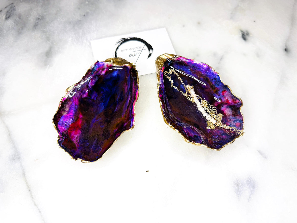 Handmade Dorset oyster shell salt and pepper pinch pot set featuring dramatic purple, blue and magenta “sea cave” interiors with hand-painted liquid gold leaf edging. Coastal ocean-inspired shell dishes with glossy resin finish and matching gold crystal spoons, styled on a white marble background.