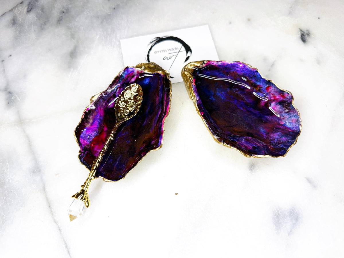 Handmade Dorset oyster shell salt and pepper pinch pot set featuring dramatic purple, blue and magenta “sea cave” interiors with hand-painted liquid gold leaf edging. Coastal ocean-inspired shell dishes with glossy resin finish and matching gold crystal spoons, styled on a white marble background.