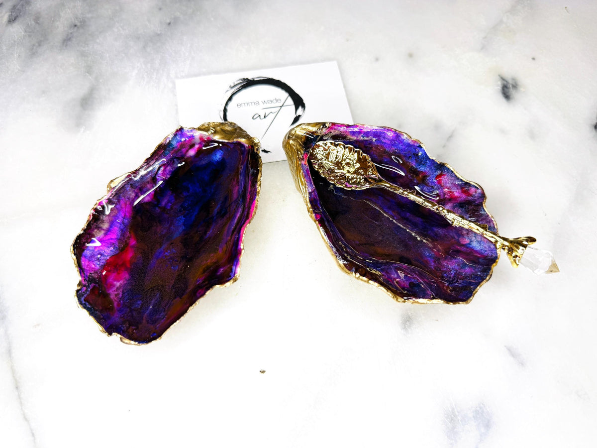 Handmade Dorset oyster shell salt and pepper pinch pot set featuring dramatic purple, blue and magenta “sea cave” interiors with hand-painted liquid gold leaf edging. Coastal ocean-inspired shell dishes with glossy resin finish and matching gold crystal spoons, styled on a white marble background.