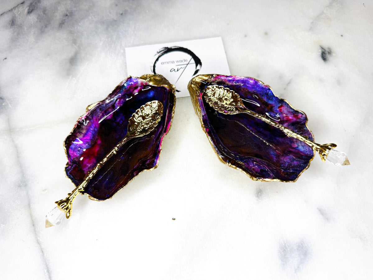 Handmade Dorset oyster shell salt and pepper pinch pot set featuring dramatic purple, blue and magenta “sea cave” interiors with hand-painted liquid gold leaf edging. Coastal ocean-inspired shell dishes with glossy resin finish and matching gold crystal spoons, styled on a white marble background.