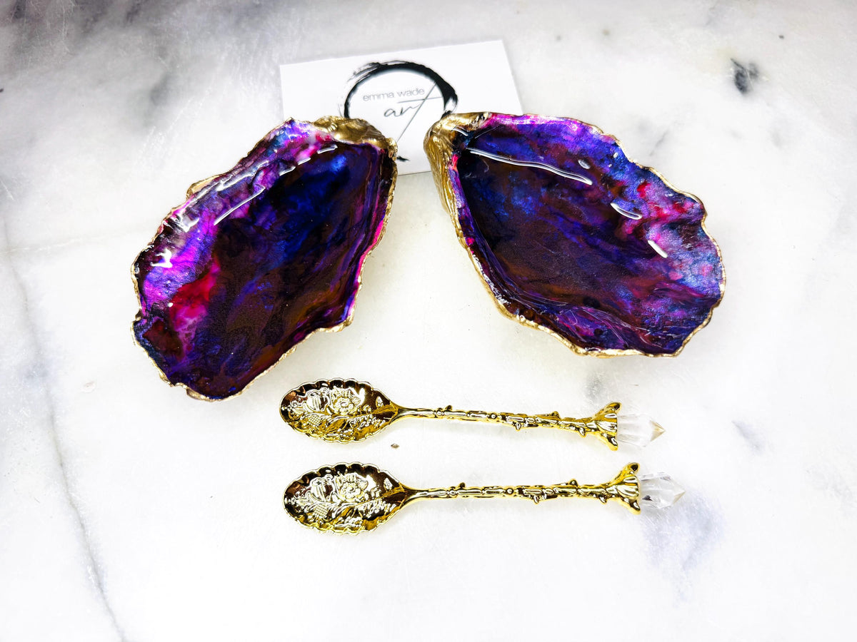 Handmade Dorset oyster shell salt and pepper pinch pot set featuring dramatic purple, blue and magenta “sea cave” interiors with hand-painted liquid gold leaf edging. Coastal ocean-inspired shell dishes with glossy resin finish and matching gold crystal spoons, styled on a white marble background.
