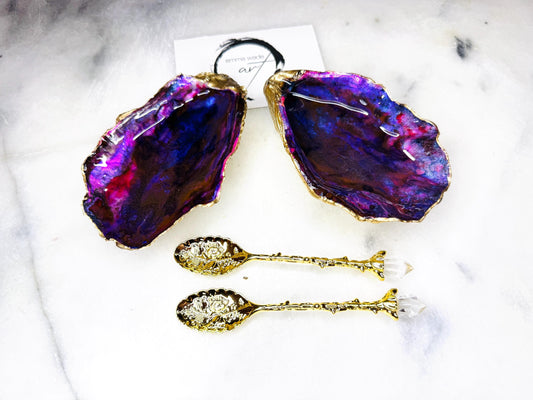 Handmade Dorset oyster shell salt and pepper pinch pot set featuring dramatic purple, blue and magenta “sea cave” interiors with hand-painted liquid gold leaf edging. Coastal ocean-inspired shell dishes with glossy resin finish and matching gold crystal spoons, styled on a white marble background.