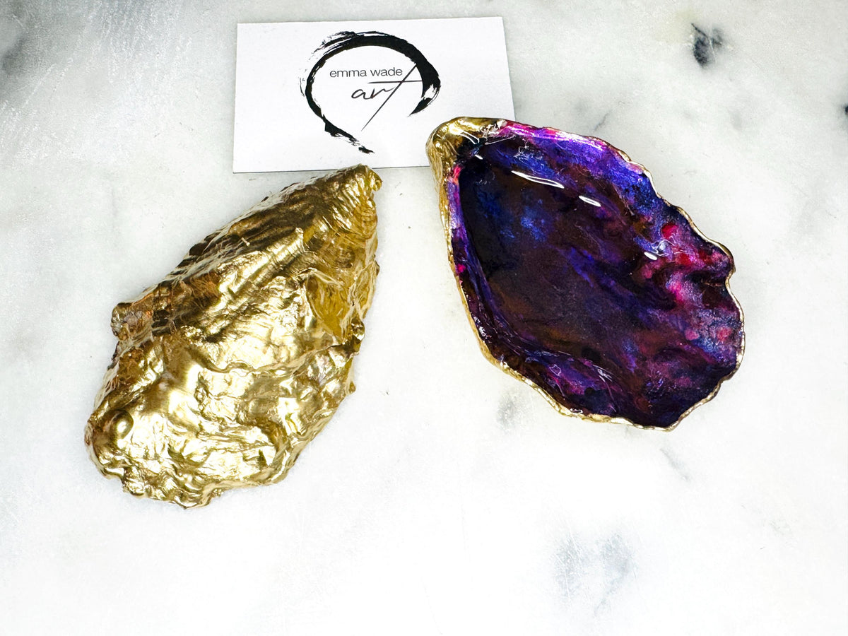 Handmade Dorset oyster shell salt and pepper pinch pot set featuring dramatic purple, blue and magenta “sea cave” interiors with hand-painted liquid gold leaf edging. Coastal ocean-inspired shell dishes with glossy resin finish and matching gold crystal spoons, styled on a white marble background.