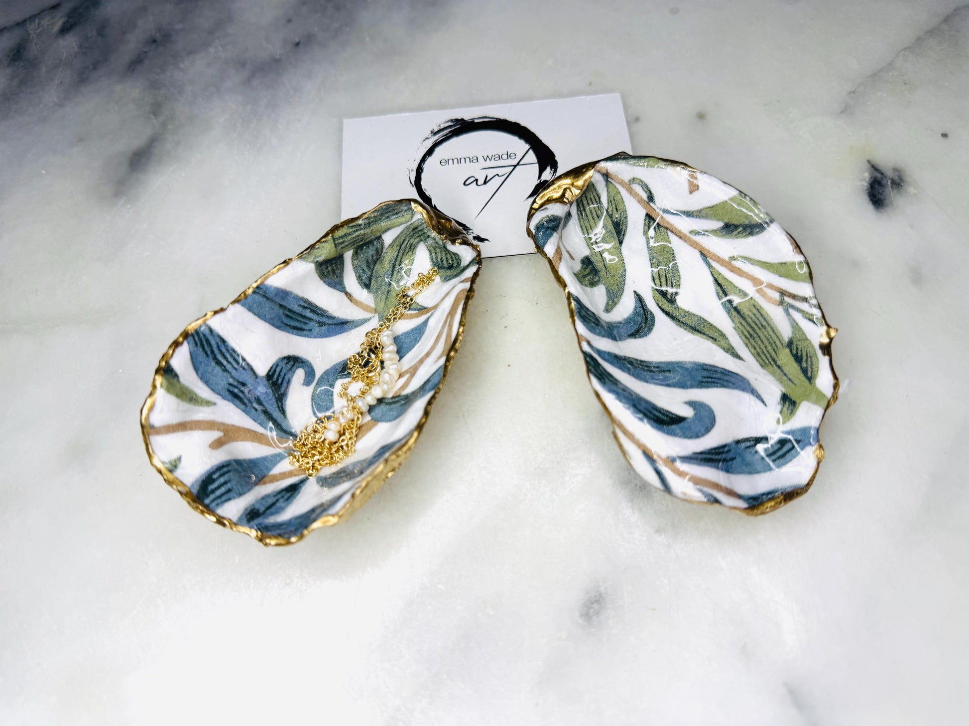 Handmade Dorset oyster shell trinket dish featuring William Morris Willow Bough botanical design in soft blue and sage green tones. Decorative jewellery holder finished with hand-applied liquid gold leaf edging and glossy resin. Coastal heritage décor shell dish styled as a ring holder and elegant bathroom accessory with natural oyster shell shape.
