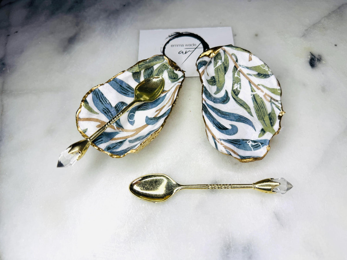 William Morris Willow Bough oyster shell salt and pepper pinch pots, sold as a pair. Natural Dorset cupped oyster shells featuring green and blue heritage pattern, hand-finished with gold leaf edging and sealed with glossy resin. Styled with optional ornate gold crystal spoons on a marble surface as elegant coastal table decor and luxury host gift.