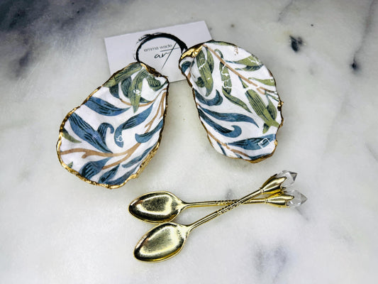 William Morris Willow Bough oyster shell salt and pepper pinch pots, sold as a pair. Natural Dorset cupped oyster shells featuring green and blue heritage pattern, hand-finished with gold leaf edging and sealed with glossy resin. Styled with optional ornate gold crystal spoons on a marble surface as elegant coastal table decor and luxury host gift.