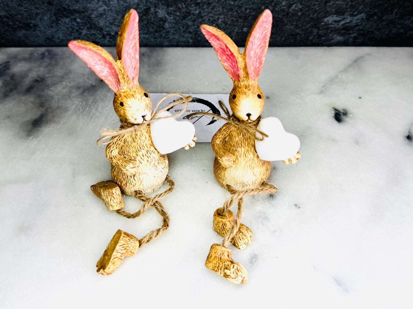 Hand finished rustic bunny shelf sitters holding white wooden hearts, gold and grey rabbit ornaments with pink ears and dangling jute legs, styled on marble surface as charming year-round home decor.