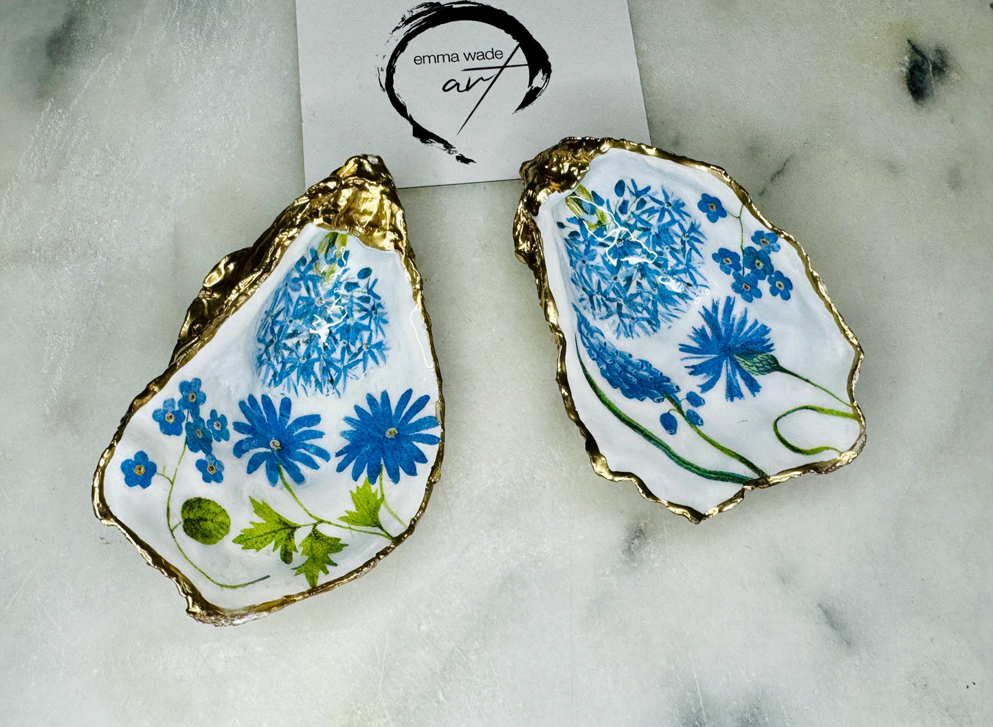 Handmade Dorset oyster shell trinket dish featuring delicate blue floral artwork including bluebells and forget-me-nots, finished with hand-applied gold leaf edging and glossy resin. Coastal-inspired jewellery dish and ring holder available in multiple sizes including XL oyster shells. Elegant handmade home décor gift ideal for birthdays, bridesmaids, Mother’s Day, weddings, and coastal interiors.