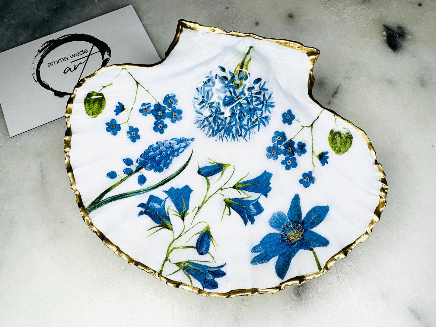 Handcrafted Dorset scallop shell trinket dish with blue spring flower design, finished with a gold leaf rim and glossy resin. The shell shows delicate blue floral motifs with green stems on a natural shell, styled on a light background. A coastal inspired jewellery or ring holder, perfect as a decorative keepsake dish, bathroom accent, or thoughtful handmade gift.