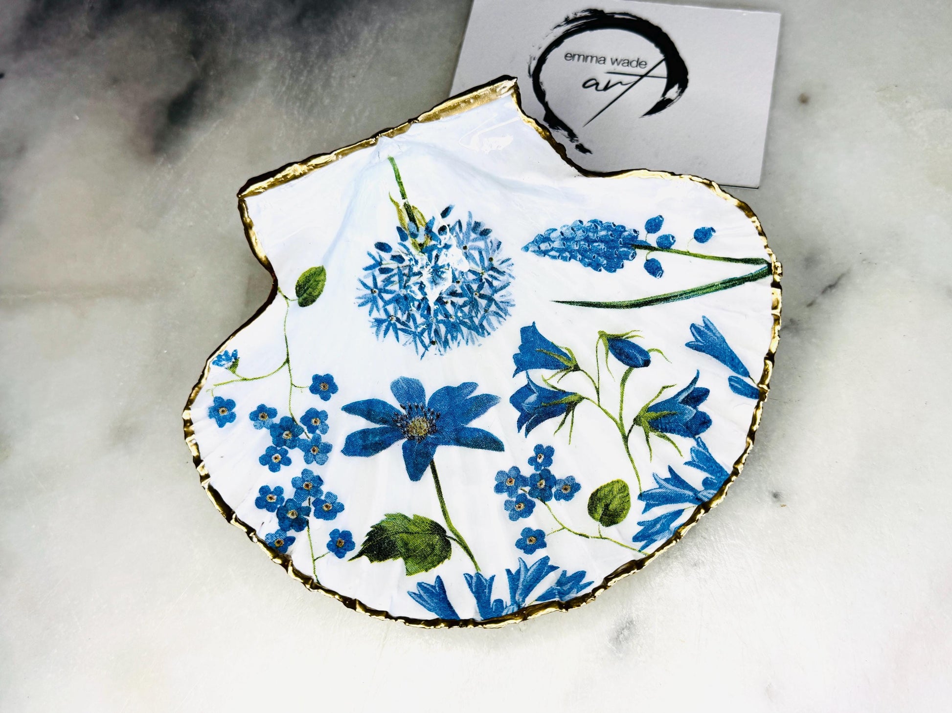 Handcrafted Dorset scallop shell trinket dish with blue spring flower design, finished with a gold leaf rim and glossy resin. The shell shows delicate blue floral motifs with green stems on a natural shell, styled on a light background. A coastal inspired jewellery or ring holder, perfect as a decorative keepsake dish, bathroom accent, or thoughtful handmade gift.