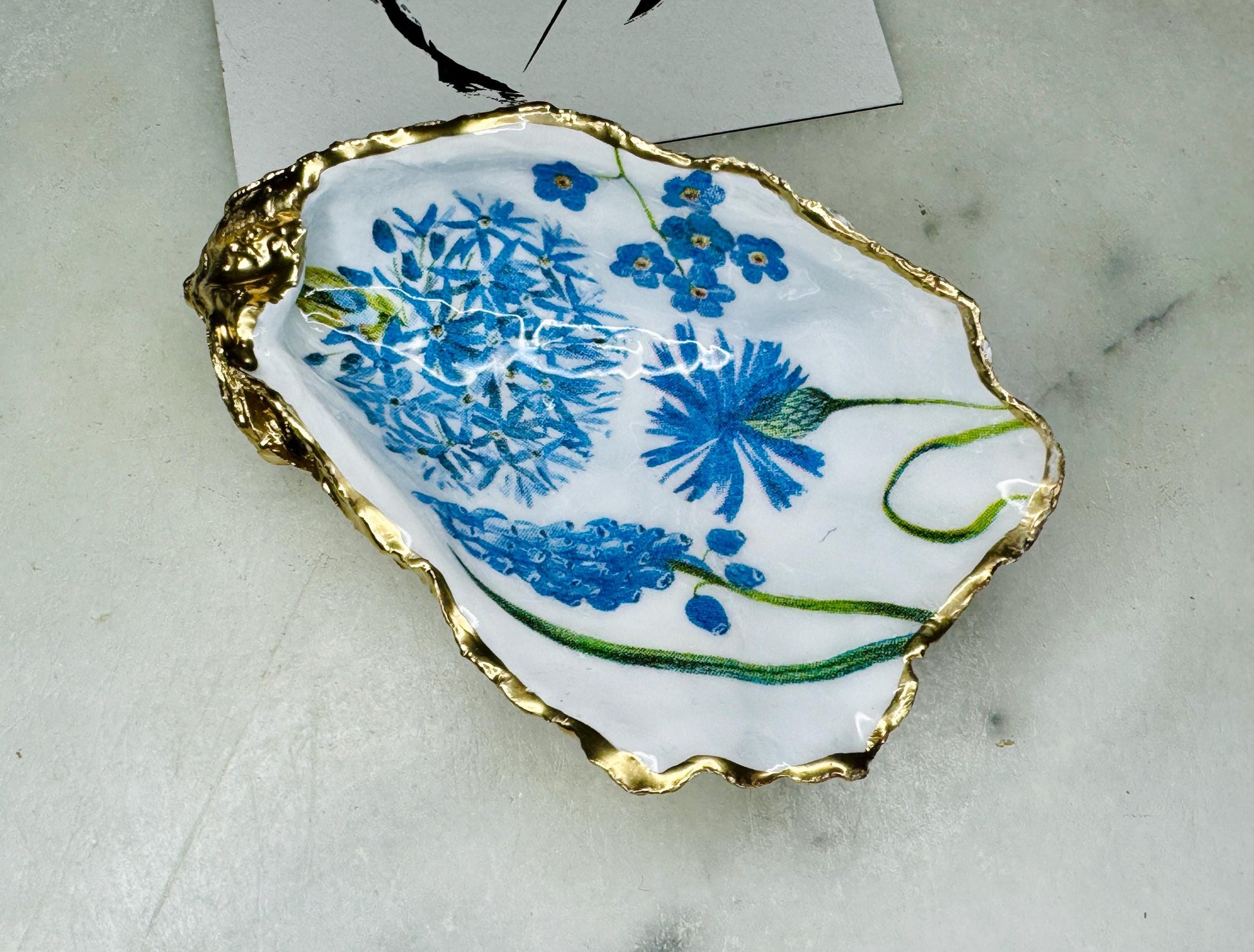 Handmade Dorset oyster shell trinket dish featuring delicate blue floral artwork including bluebells and forget-me-nots, finished with hand-applied gold leaf edging and glossy resin. Coastal-inspired jewellery dish and ring holder available in multiple sizes including XL oyster shells. Elegant handmade home décor gift ideal for birthdays, bridesmaids, Mother’s Day, weddings, and coastal interiors.