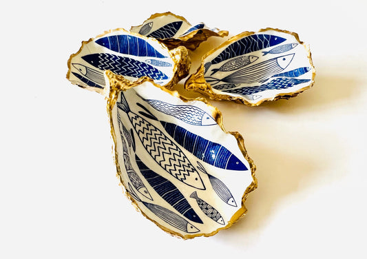 Luxury hand gilded Dorset oyster shell trinket dish featuring a refined blue fish motif, edged in liquid gold leaf and sealed in crystal clear resin. Sustainable coastal ring holder handmade by the artist in Surrey and presented in eco friendly branded gift packaging. Elegant beach house decor piece ideal for jewellery storage, wedding styling or sophisticated coastal interiors.