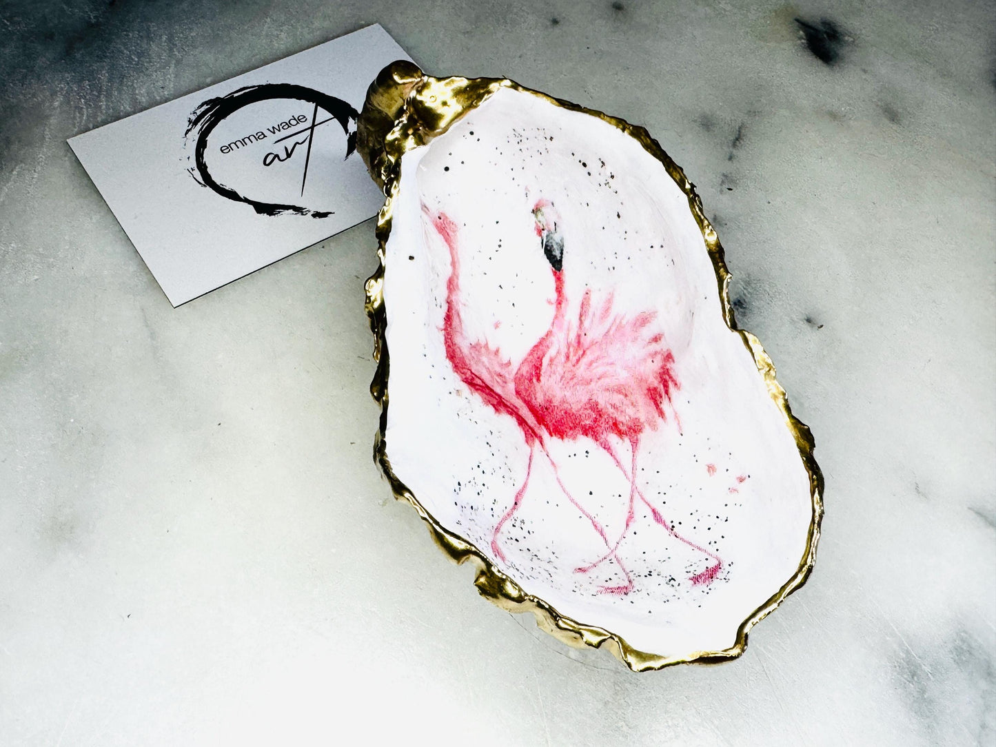 Handmade Dorset oyster shell trinket dishes featuring elegant pink flamingo bird artwork with hand-painted gold leaf edging and glossy crystal clear resin. Coastal flamingo jewellery holders styled on a white marble background with tropical blush pink tones and luxury coastal home décor styling.