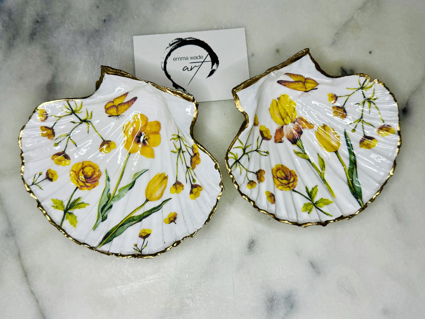 Hand finished scallop shell trinket dish featuring yellow spring flower artwork with a hand gilded gold leaf rim, sealed in glossy clear resin for durability and shine. Coastal inspired shell jewellery dish photographed on a light surface, ideal for rings, earrings and small keepsakes. Decorative floral shell dish by EmmaWadeART inspired by the Dorset coastline, perfect as a gift or elegant home décor accent.