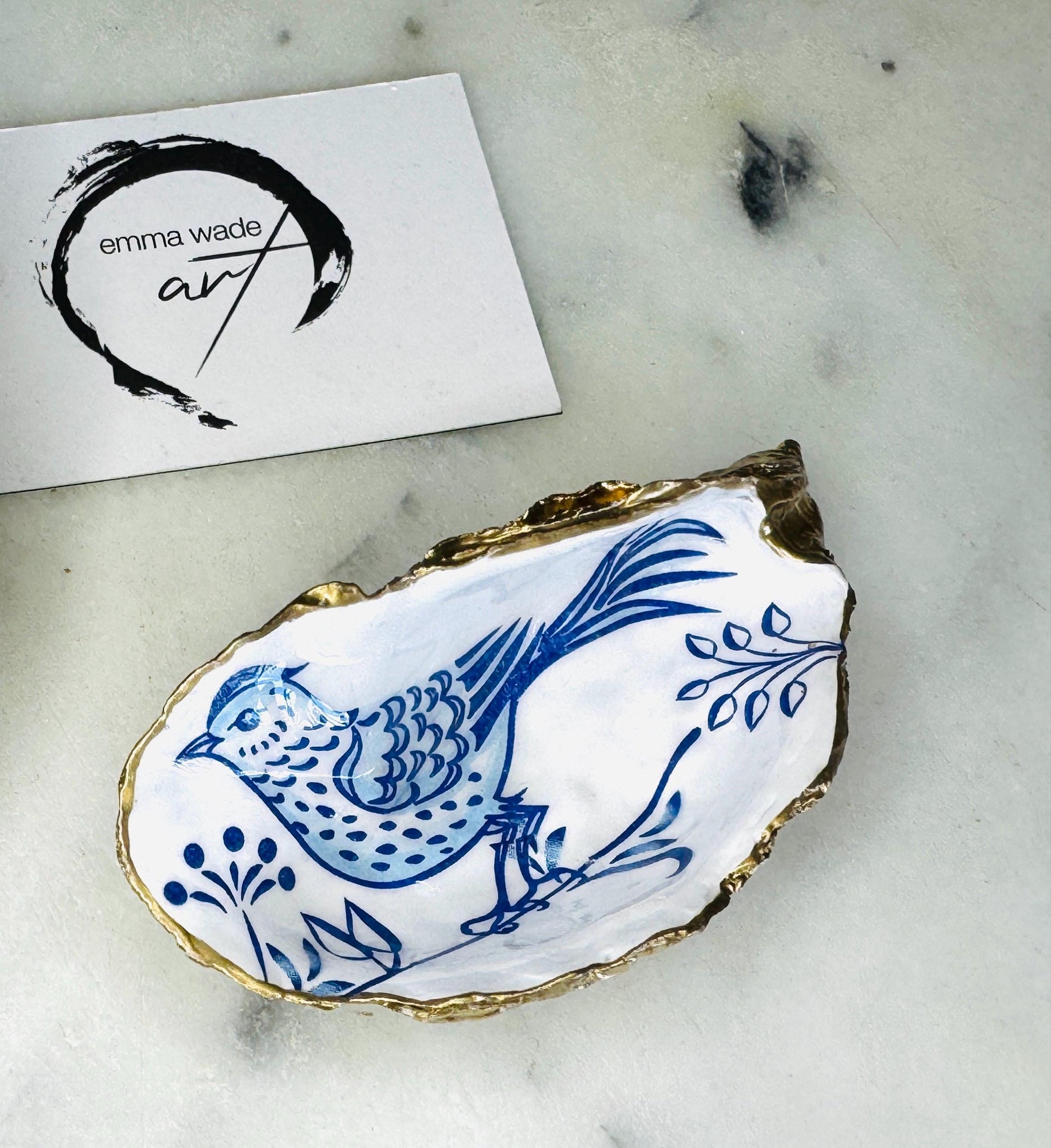 Handmade Dorset oyster shell trinket dish with blue porcelain-style bird design, gold leaf gilded edges and glossy resin finish. Coastal jewellery holder and ring dish photographed on a light surface, showing the natural shell shape and pearly interior. Unique sustainably sourced shell, sold individually as decorative coastal home décor or a thoughtful gift.