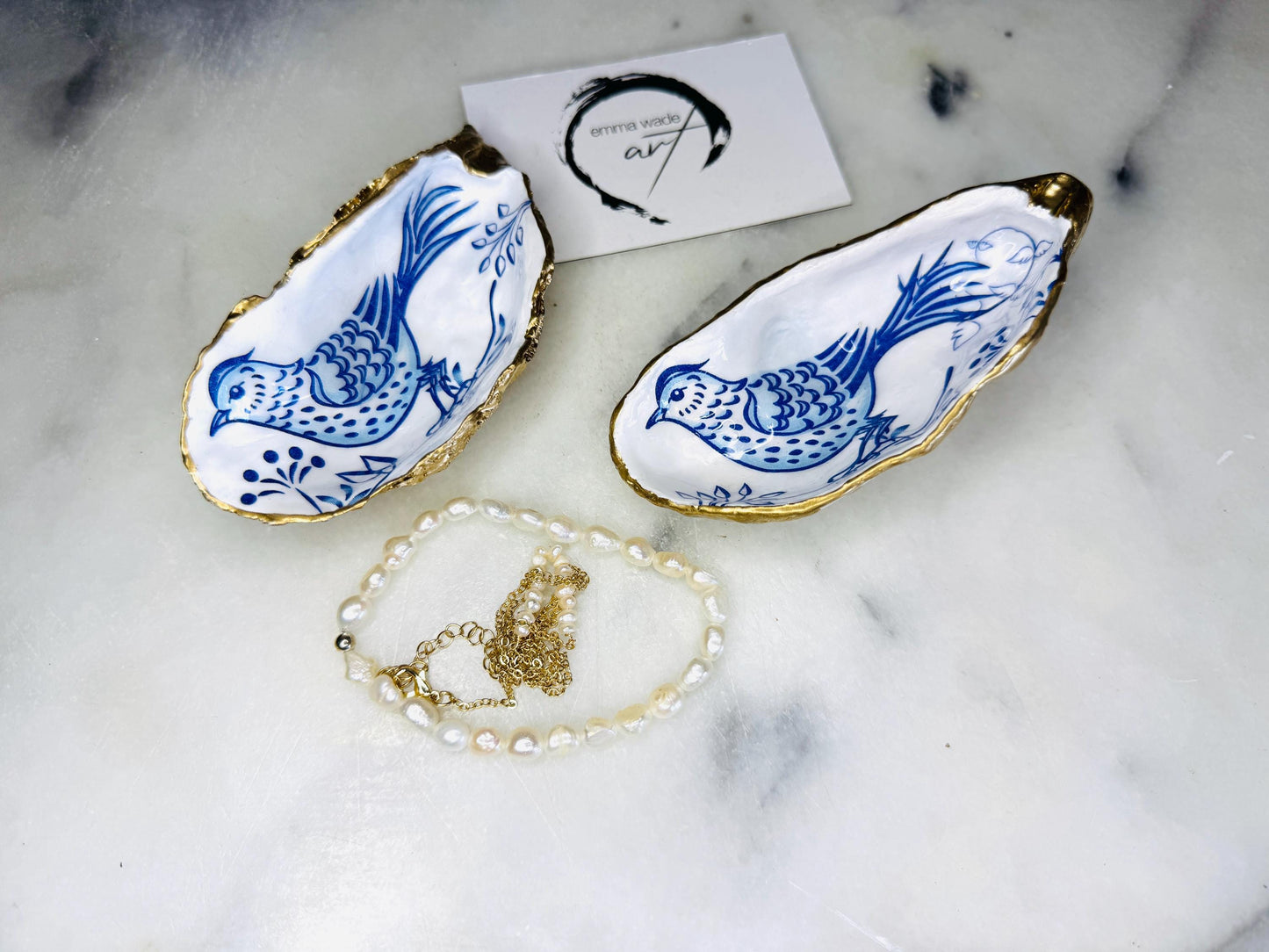 Handmade Dorset oyster shell trinket dish with blue porcelain-style bird design, gold leaf gilded edges and glossy resin finish. Coastal jewellery holder and ring dish photographed on a light surface, showing the natural shell shape and pearly interior. Unique sustainably sourced shell, sold individually as decorative coastal home décor or a thoughtful gift.