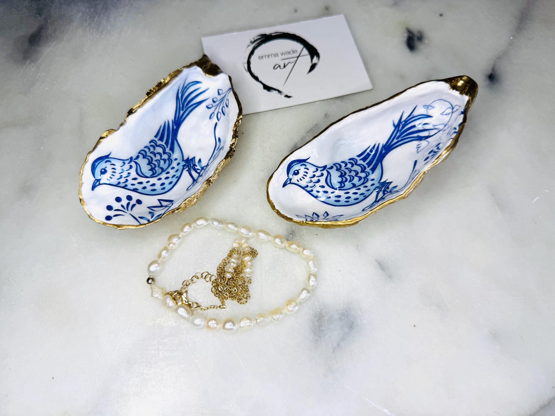 Handmade Dorset oyster shell trinket dish with blue porcelain-style bird design, gold leaf gilded edges and glossy resin finish. Coastal jewellery holder and ring dish photographed on a light surface, showing the natural shell shape and pearly interior. Unique sustainably sourced shell, sold individually as decorative coastal home décor or a thoughtful gift.