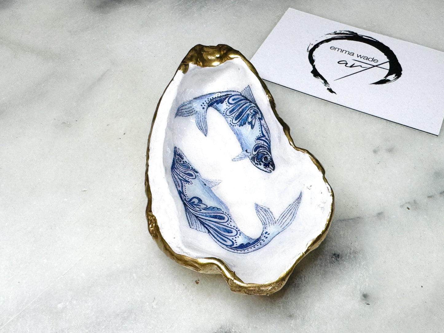 Handmade Dorset oyster shell trinket dish with blue and white paisley fish design, gold leaf gilded edges and glossy resin finish. Coastal jewellery holder and ring dish, photographed on a light surface showing the natural shell shape and pearly interior. Unique sustainably sourced shell, sold individually as a decorative coastal home décor accent or gift.