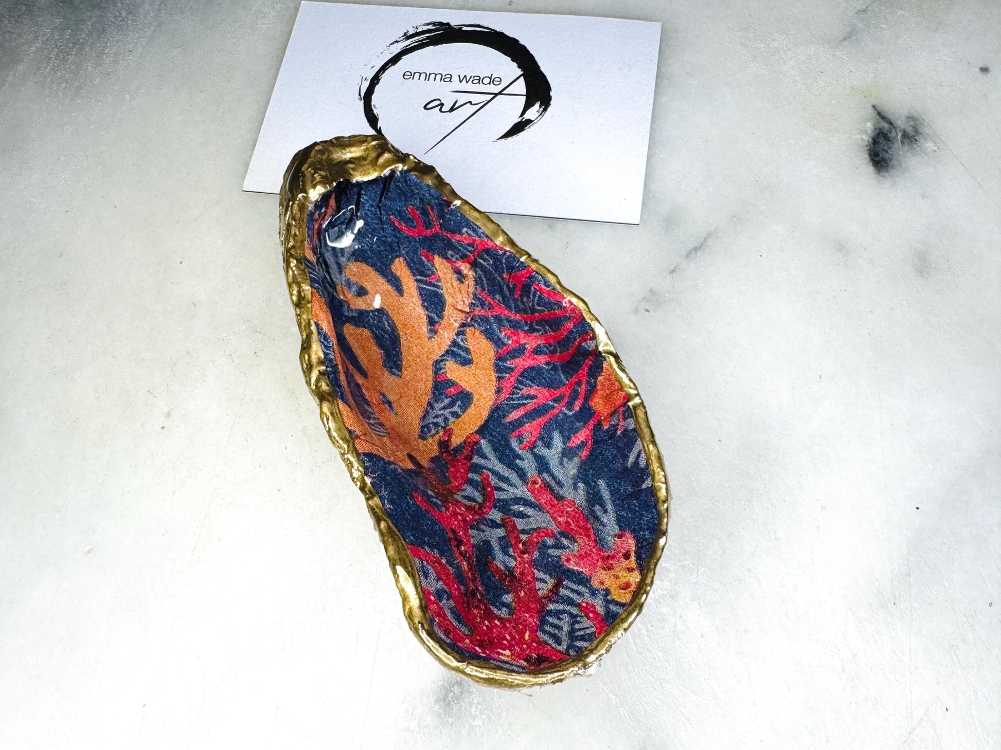 Handmade Dorset oyster shell trinket dishes featuring vibrant sea coral inspired artwork in coral red, navy and turquoise tones, finished with hand-painted gold leaf edging and glossy crystal clear resin. Coastal jewellery dishes styled on a white marble background with rich ocean-inspired botanical patterns and luxurious gilded edges.