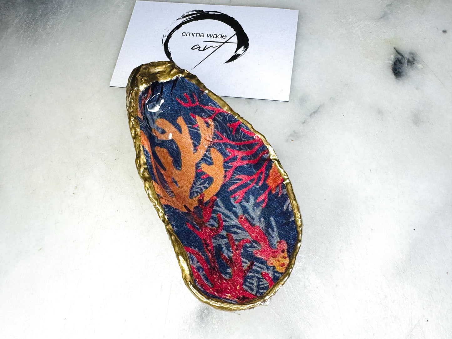 Handmade Dorset oyster shell trinket dishes featuring vibrant sea coral inspired artwork in coral red, navy and turquoise tones, finished with hand-painted gold leaf edging and glossy crystal clear resin. Coastal jewellery dishes styled on a white marble background with rich ocean-inspired botanical patterns and luxurious gilded edges.