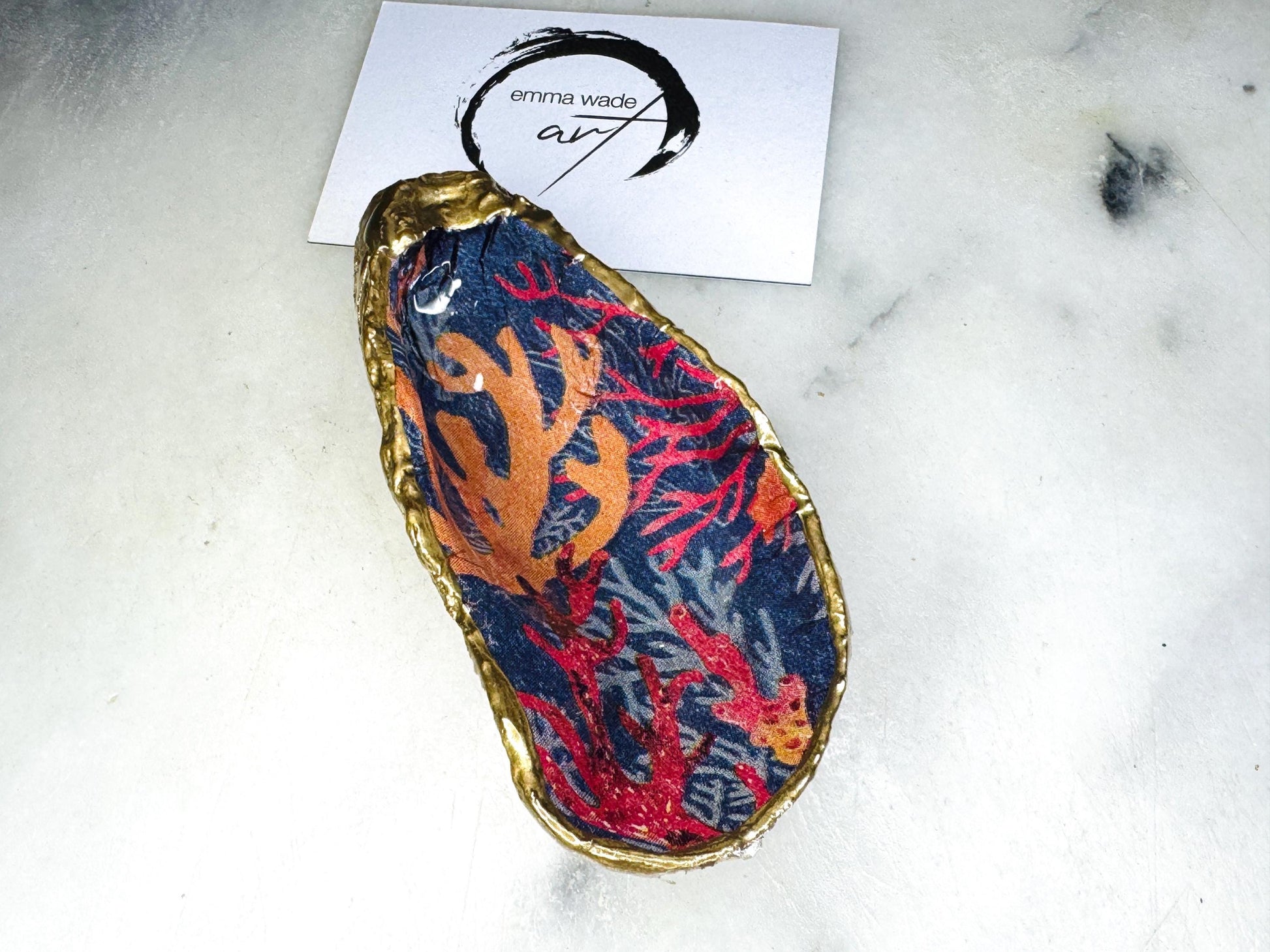 Handmade Dorset oyster shell trinket dishes featuring vibrant sea coral inspired artwork in coral red, navy and turquoise tones, finished with hand-painted gold leaf edging and glossy crystal clear resin. Coastal jewellery dishes styled on a white marble background with rich ocean-inspired botanical patterns and luxurious gilded edges.